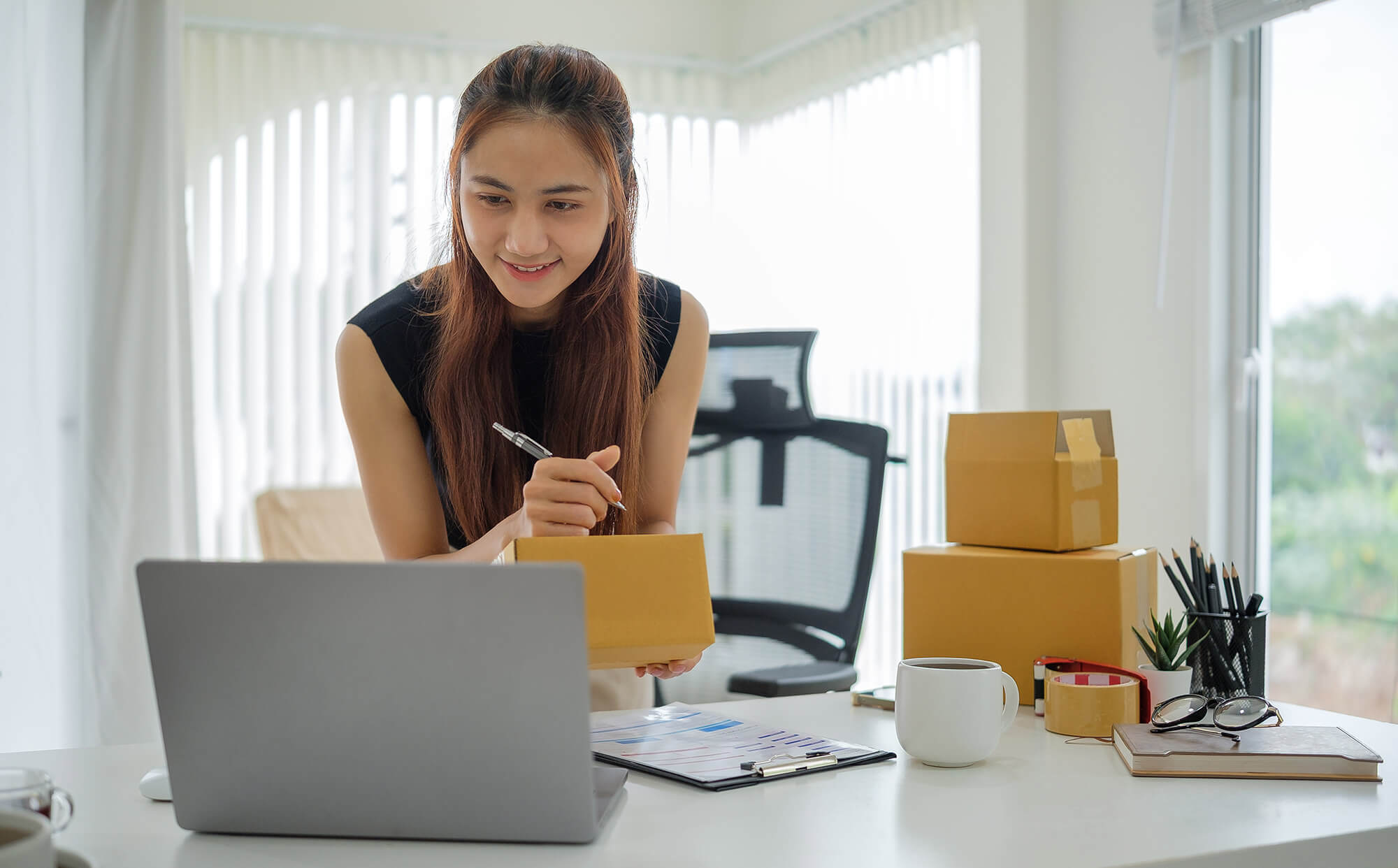 An ecommerce business owner prepares Shopify orders at her desk, holding a shipping box while looking at her laptop. Several other packages, a clipboard with charts, and office supplies are visible, showing her managing her online store.