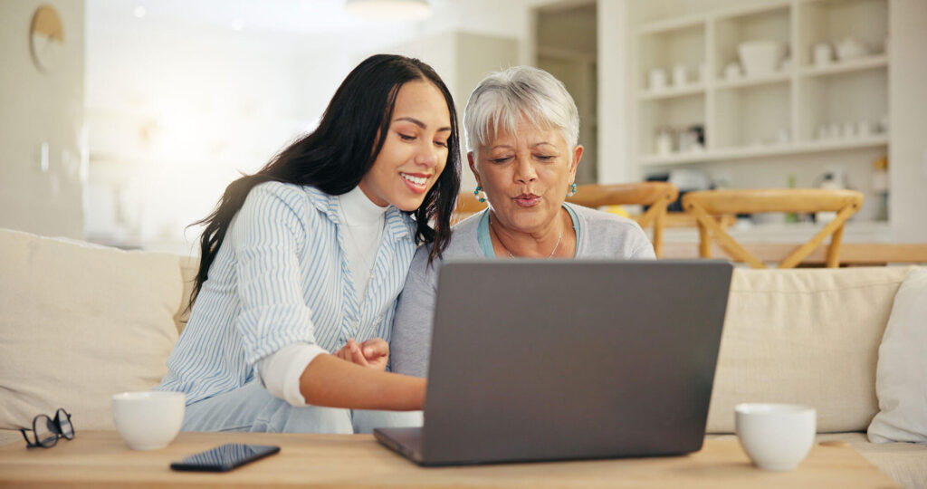 A younger woman and an older woman sit together on a couch, smiling as they look at a laptop screen in a bright, cozy living room.