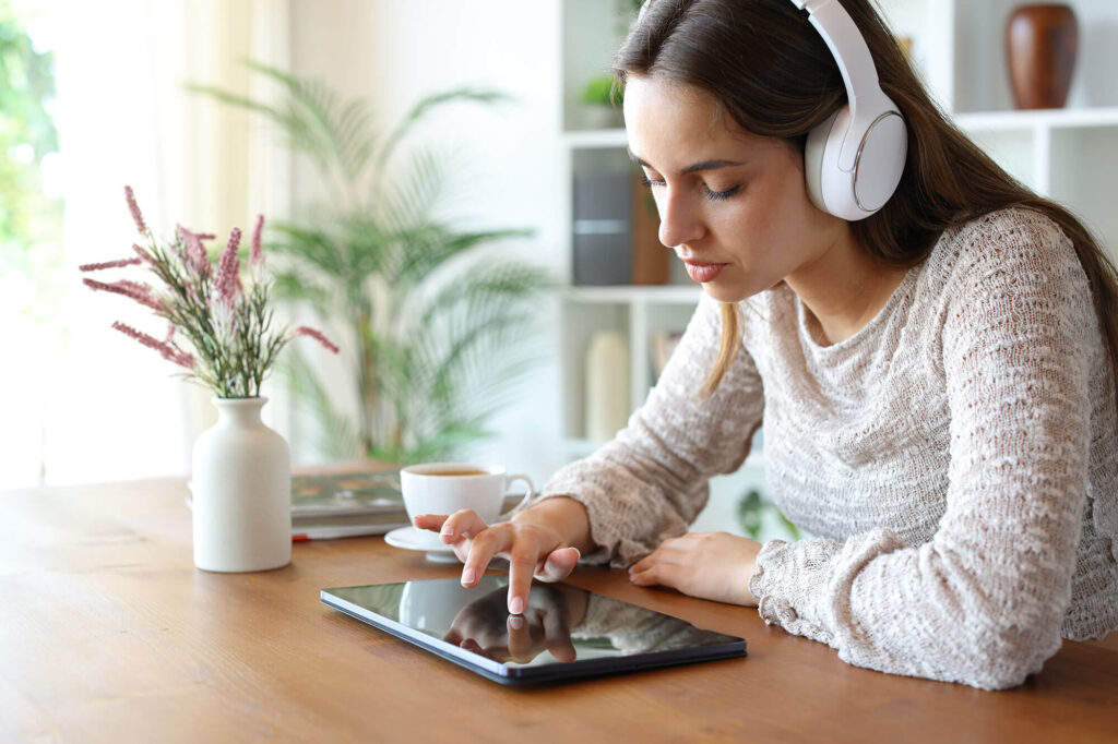 A woman wearing headphones sits at a wooden table, using a tablet with her finger while a cup of coffee and a vase of flowers sit nearby.