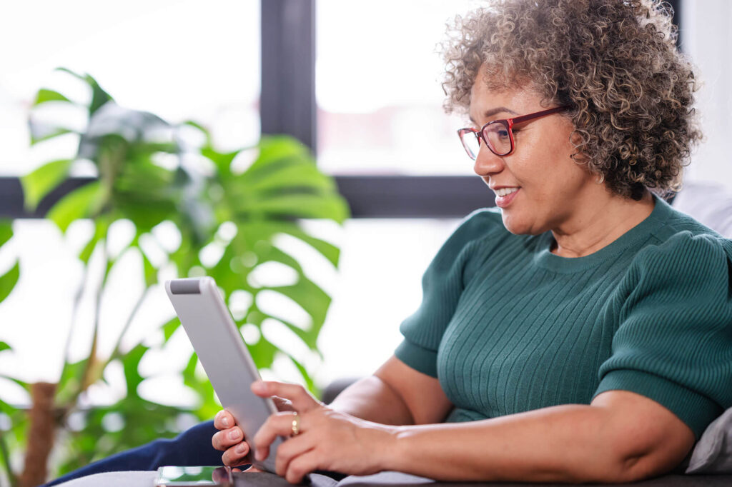 A woman wearing glasses and a green sweater smiles while using a tablet, sitting near a window with leafy green plants in the background.