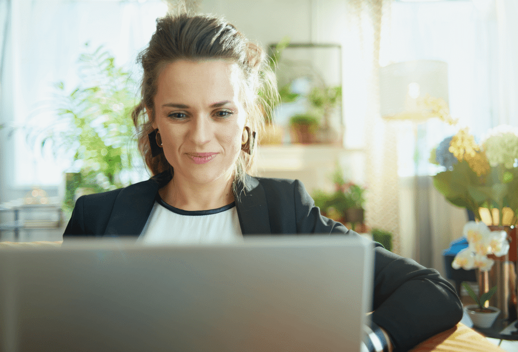 A woman smiling while working on a laptop in a bright home office filled with plants and natural light.